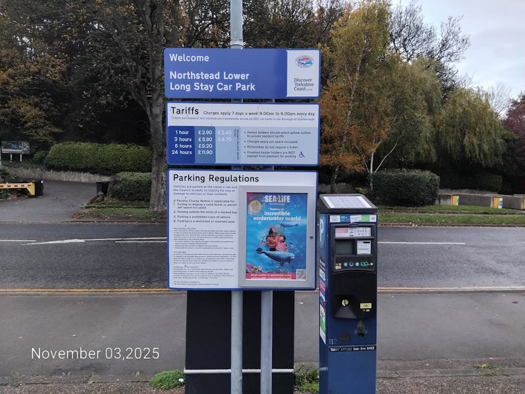 Sign for Northstead Lower Long Stay Car Park with parking tariffs and regulations, near trees and a pay machine.