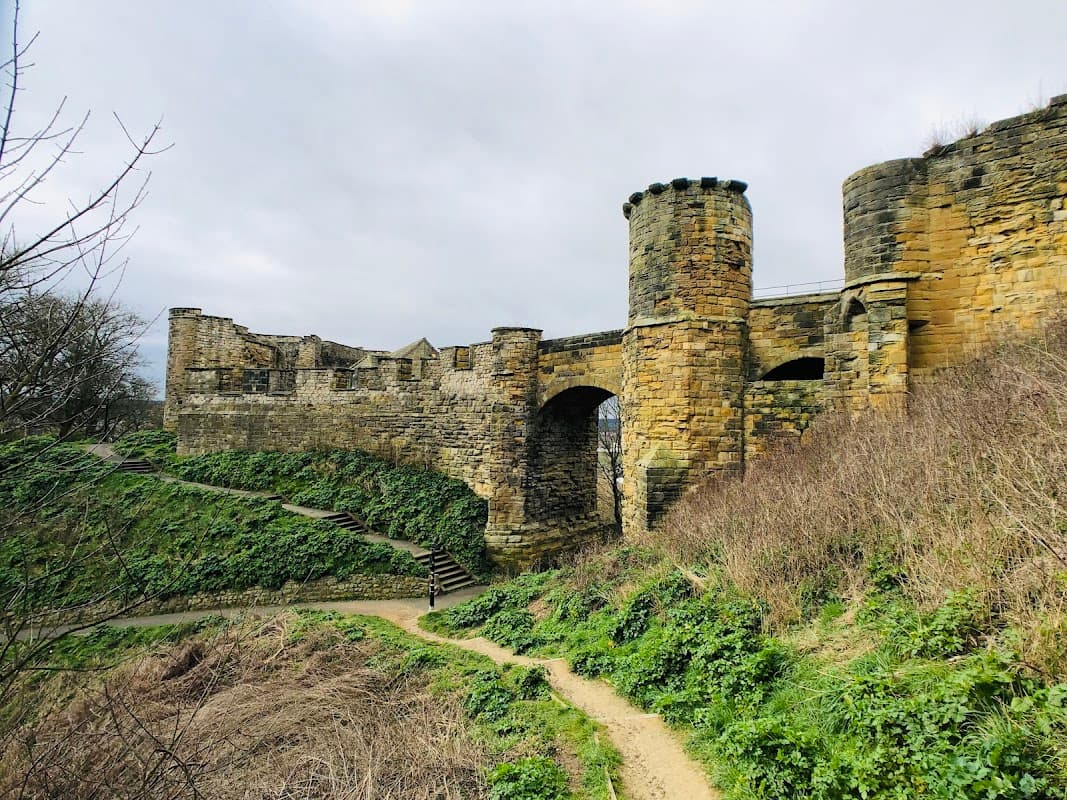 Stone castle ruins with towers, surrounded by greenery and a winding path in a cloudy sky.