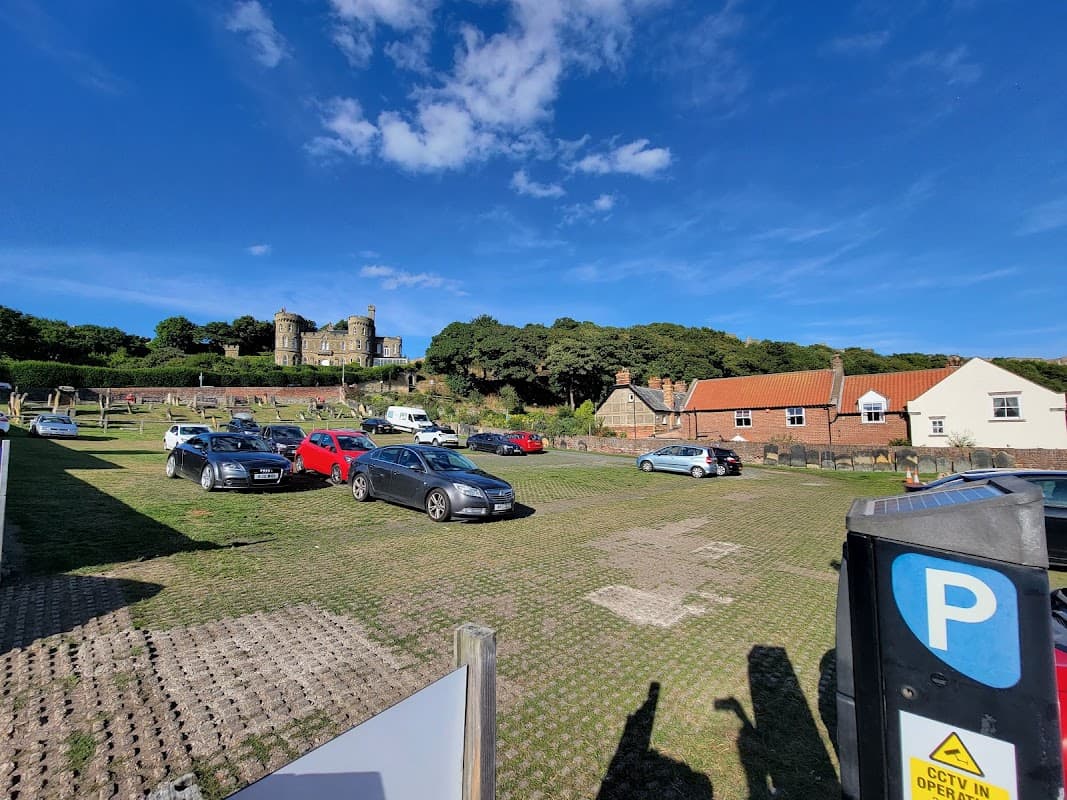 St Mary's pay and display car park with parked cars, green grass, and a historic building on a hill under a blue sky.