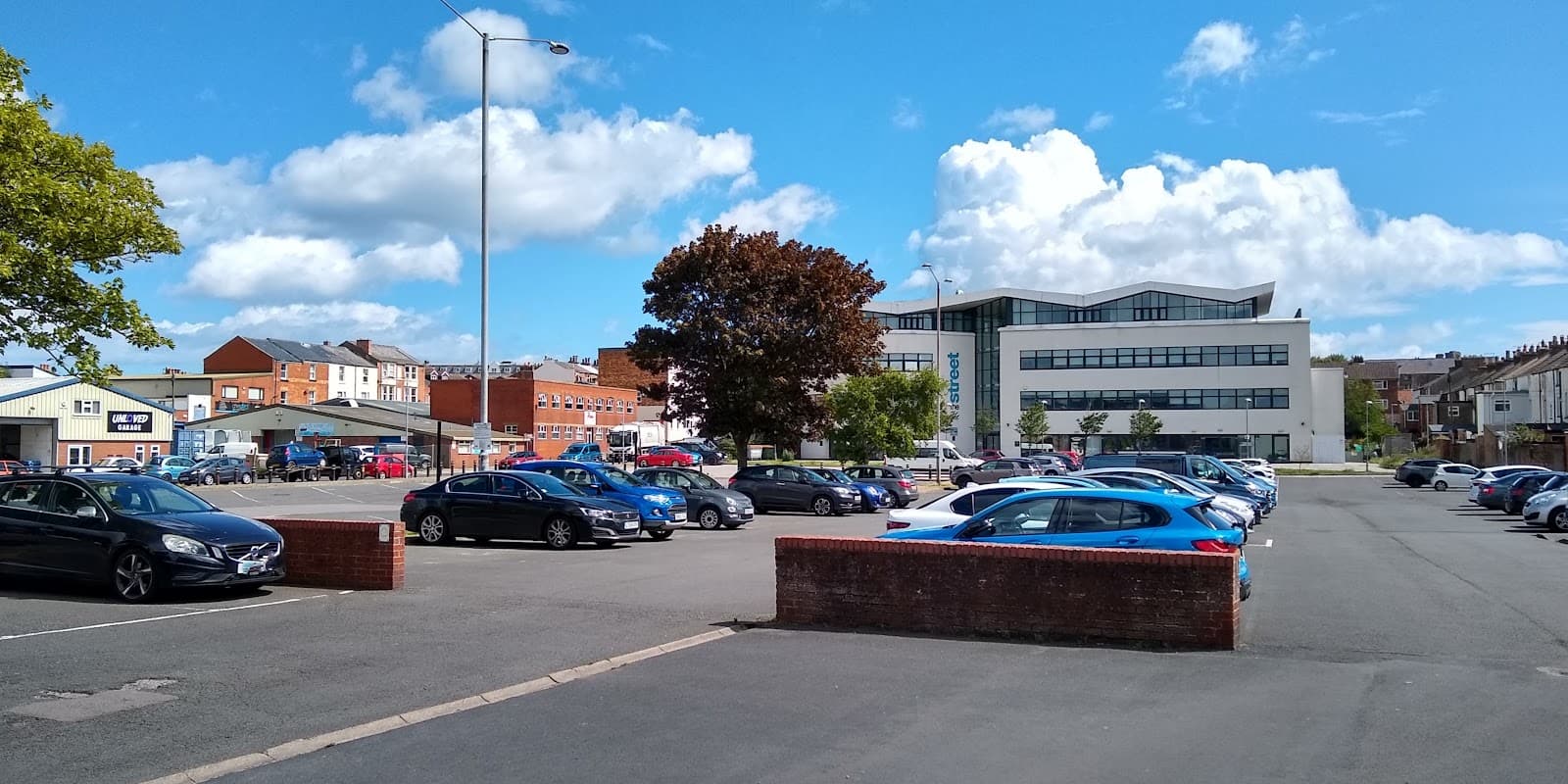 Spacious car park with various parked cars, trees, and a modern building under a blue sky with scattered clouds.