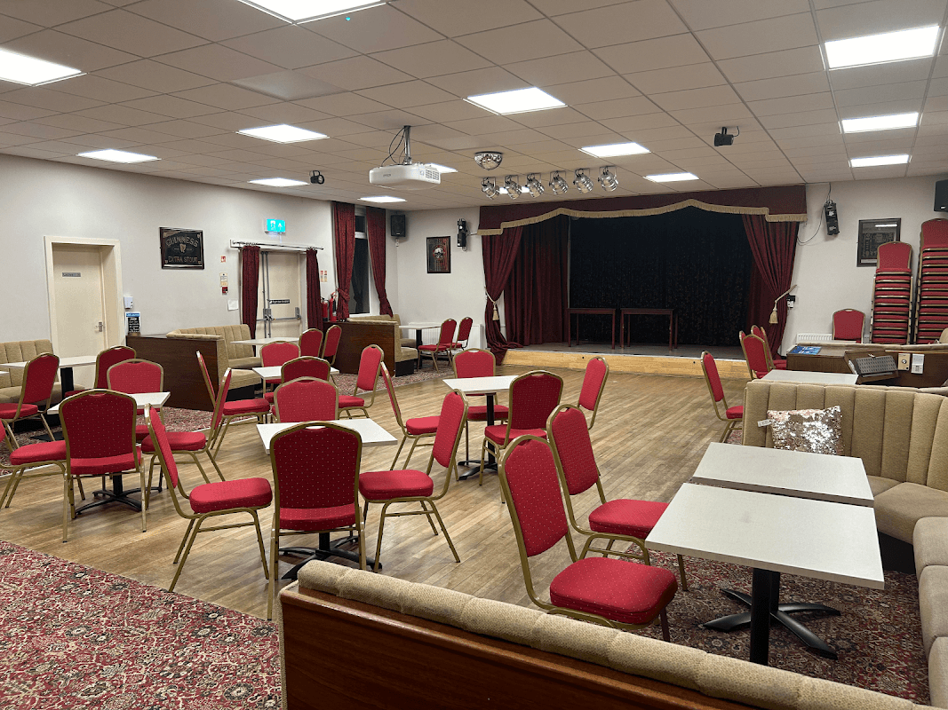 Interior of Scissett Club Ltd with red chairs, tables, a stage, and a carpeted floor in a cozy setting.