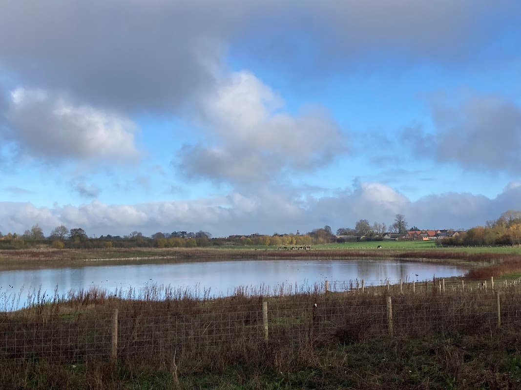 Serene lake surrounded by lush greenery and distant houses under a partly cloudy sky in Scorton, North Yorkshire.