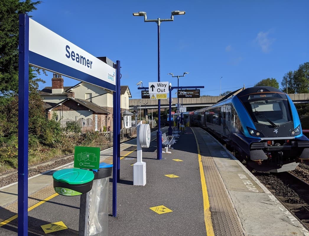Seamer train station platform with signage, a train, and a green hand sanitizer station under a clear blue sky.