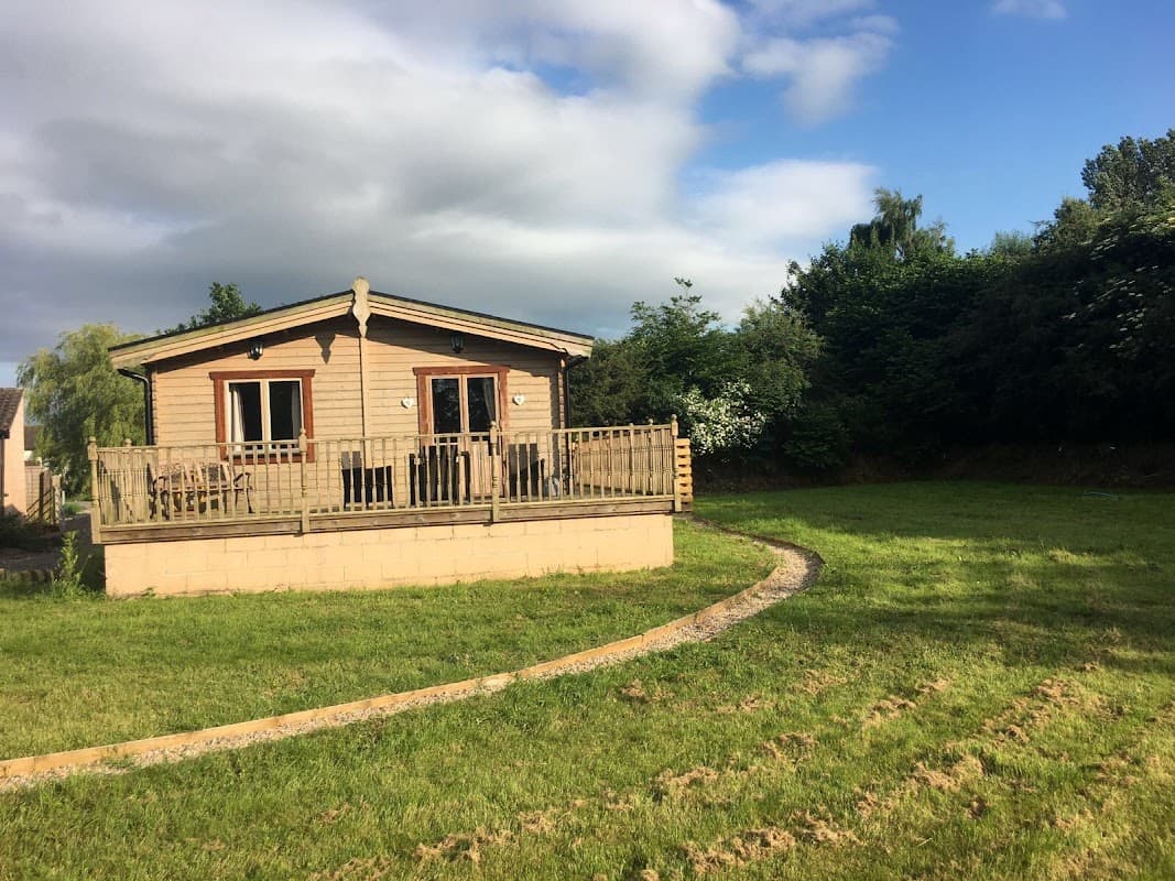 Wooden cabin with a deck, surrounded by green grass and trees under a partly cloudy sky in Seaton Ross, Yorkshire.