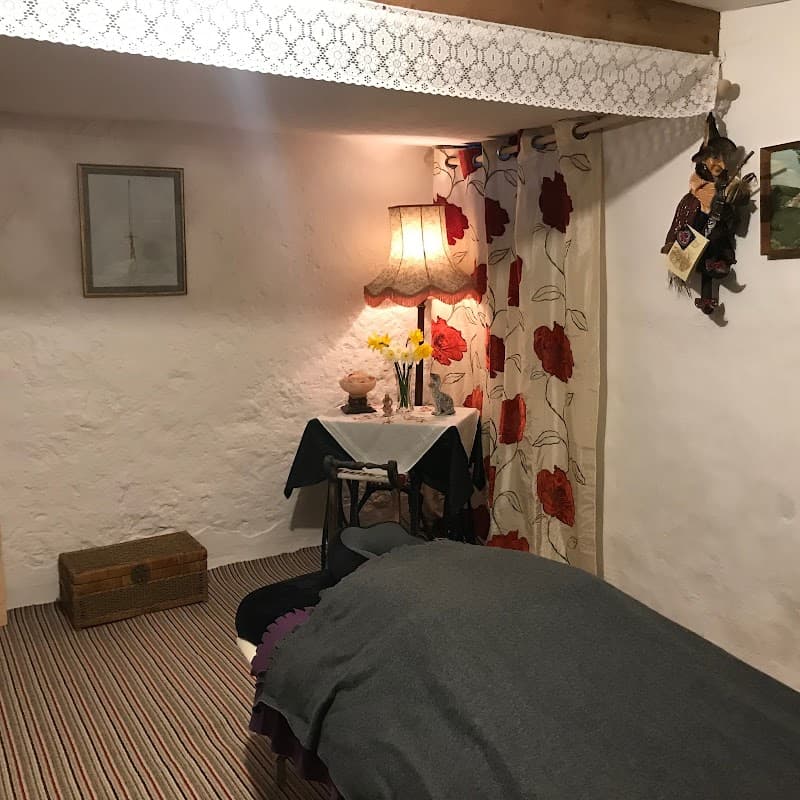 Cozy interior with a bed, floral curtains, a lamp, and a small table with flowers and decor in a rustic setting.