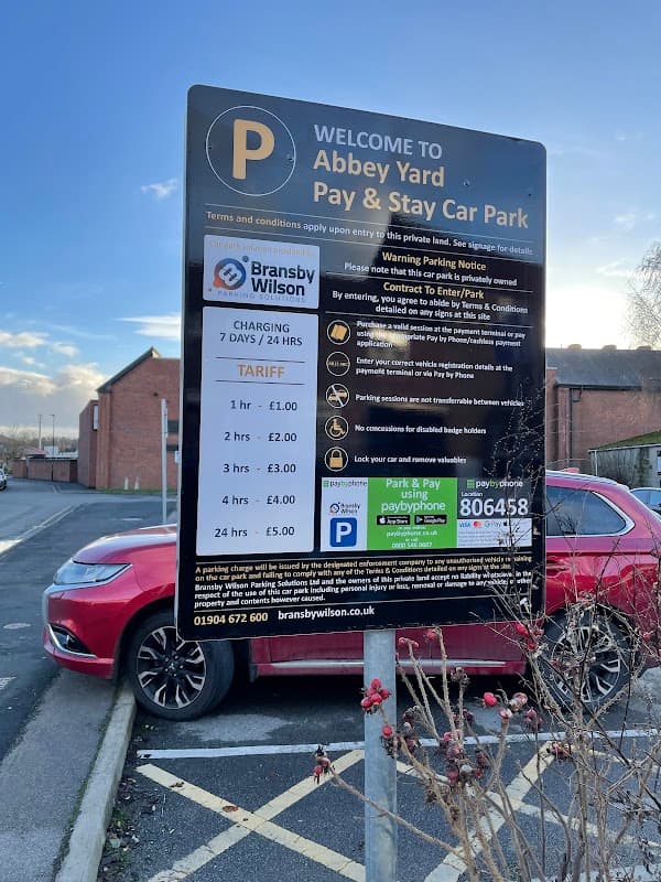 Welcome sign for Abbey Yard Pay & Display Car Park, detailing charges and contact information, with a parked car nearby.