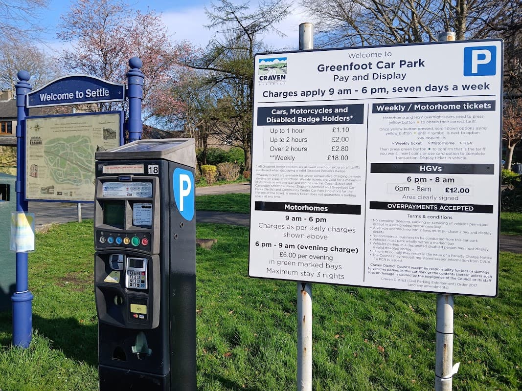 Greenfoot Car Park sign with parking rates, payment machine, and a welcome sign for Settle, surrounded by trees.