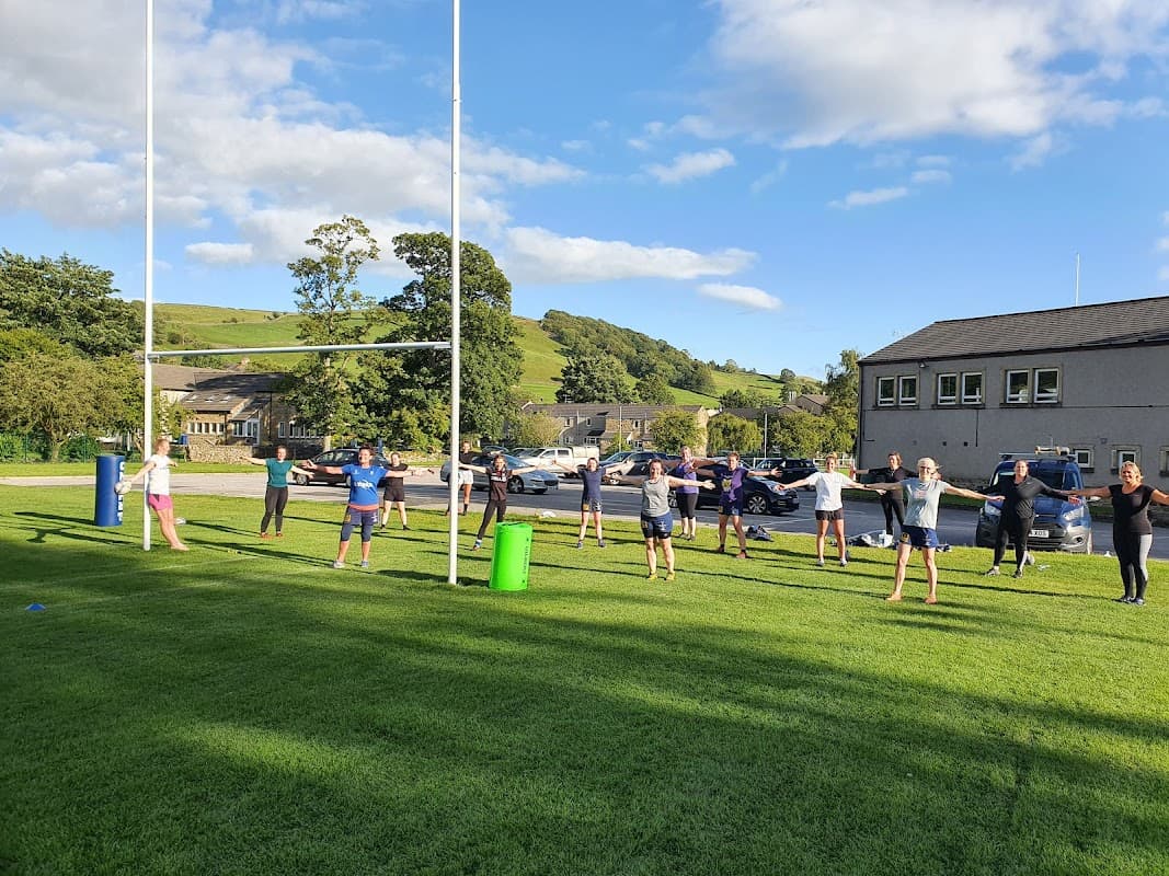 Group of people exercising on a rugby field with goalposts, surrounded by green hills and parked cars.