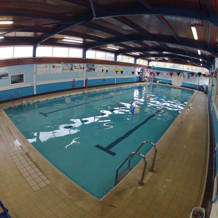 Indoor swimming pool with clear water, colorful beach balls, and safety equipment along the sides. Brightly lit area.