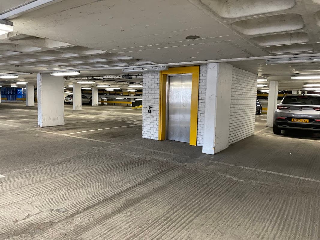 Elevator entrance with yellow door in a spacious car park, parked cars visible in the background.