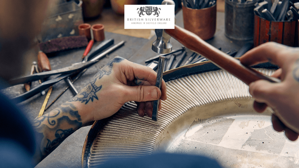 Craftsman working on intricate silverware with tools and materials scattered on a workbench. Logo at the top.