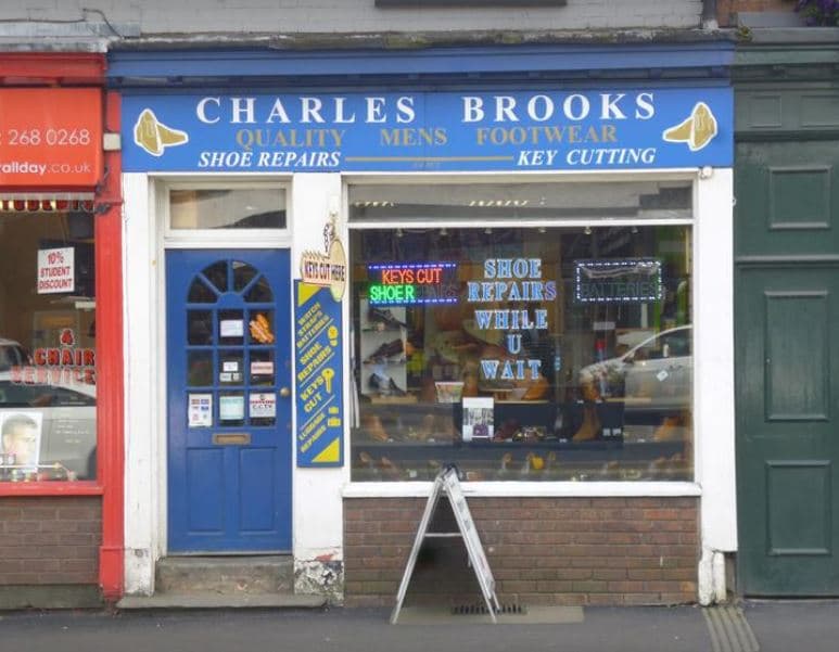 Blue storefront with "Charles Brooks" sign, display of shoes, and neon signs for shoe repairs and key cutting.