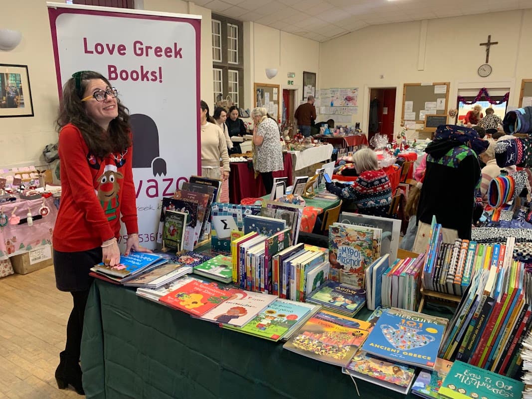 A woman in a red sweater smiles at a table filled with colorful Greek books at a bustling event in Sheffield.