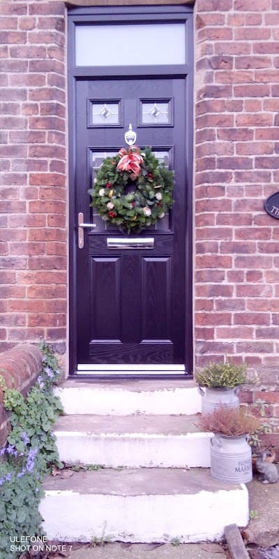 Black front door adorned with a festive wreath, surrounded by brick walls and potted plants on stone steps.