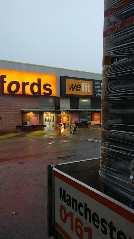 Halfords store entrance with bright signage, wet pavement, and stacked pallets in foreground.