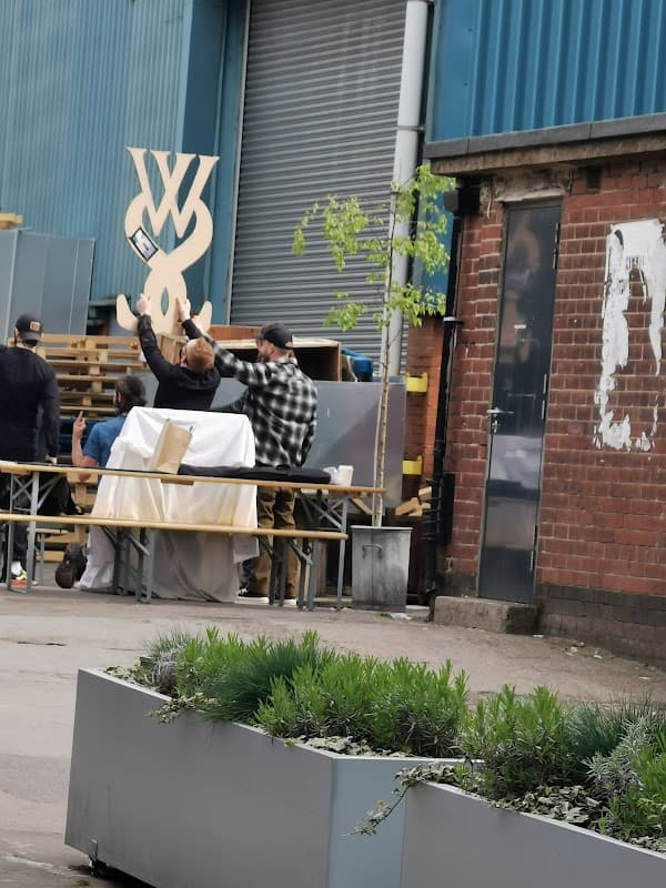 People interacting at outdoor seating with a large wooden sign in the background, surrounded by greenery and industrial buildings.