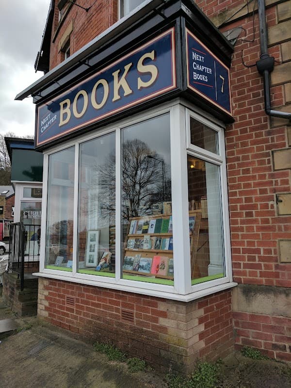 Bookshop storefront with large sign "Next Chapter Books" and display of books in the window. Red brick building.