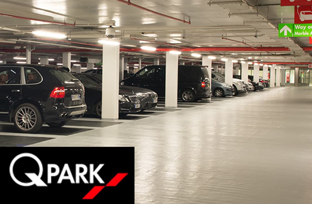 Multi-storey car park interior with parked cars, bright lighting, and directional signage for pedestrian access.