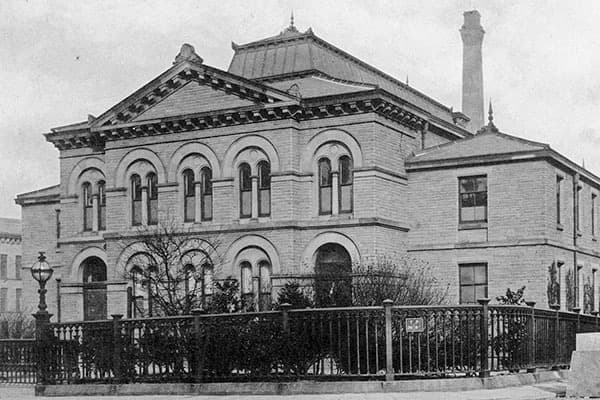 Historic building with arched windows, decorative stonework, and a prominent chimney, surrounded by greenery and a fence.