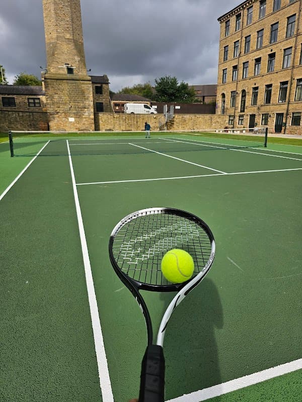 Tennis racket holding a ball, with green courts and historic stone buildings in the background under a cloudy sky.