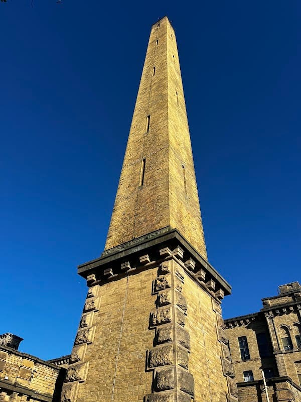 Tall stone tower against a clear blue sky, part of Salts Mill with nearby historic buildings. Free parking area visible.