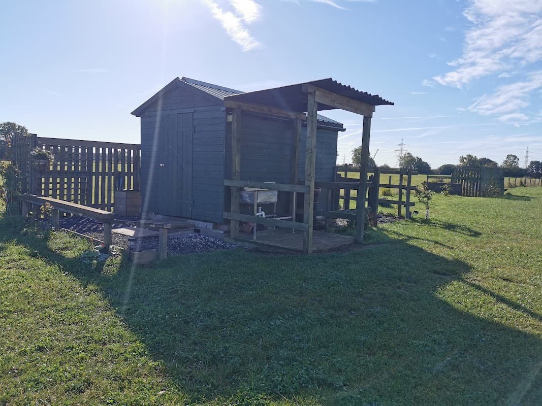 A wooden shed with a porch in a grassy field under a clear blue sky, surrounded by a wooden fence.