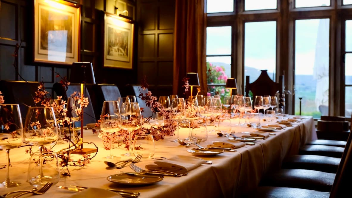 Elegant dining setup with a long table, glassware, and floral decorations, set against large windows in a cozy restaurant.