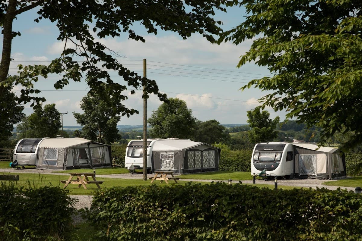 Caravans set in a lush green landscape, surrounded by trees and distant hills under a clear blue sky.