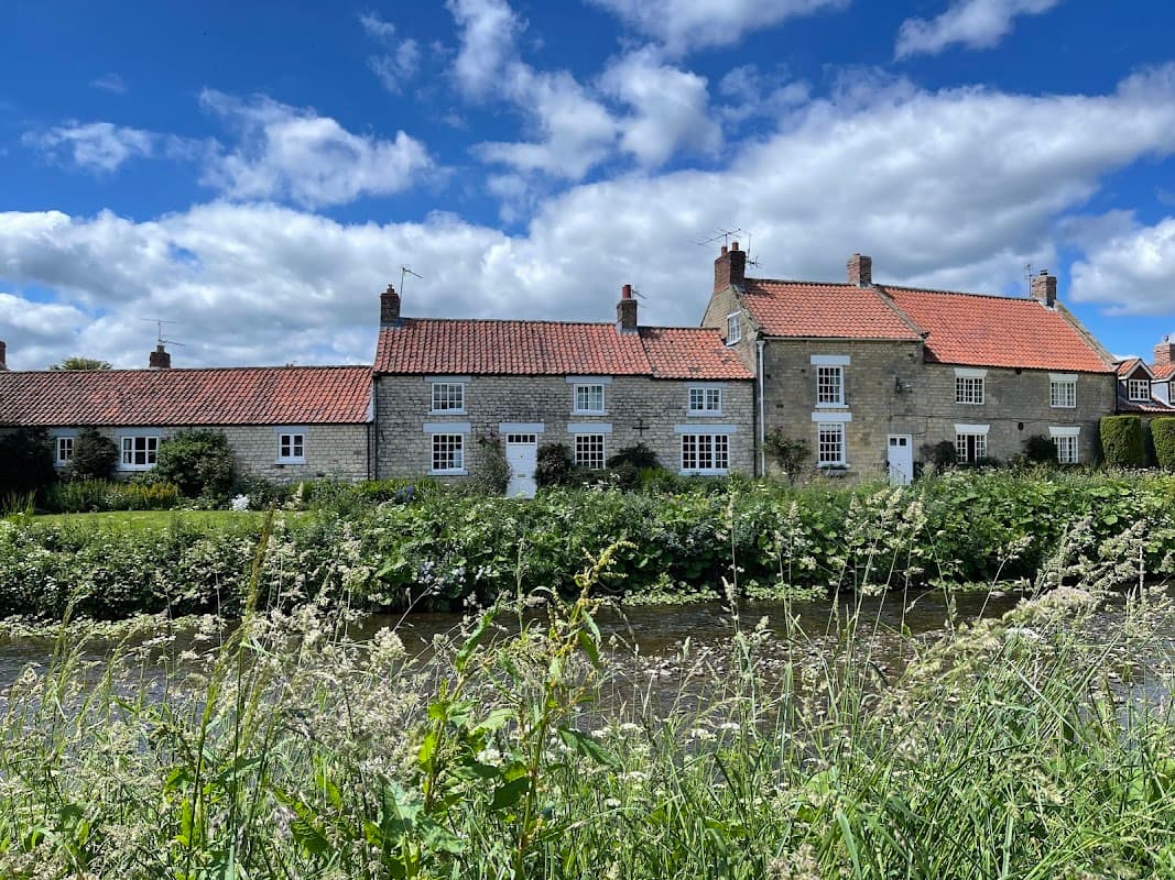 The Fox & Hounds Country Inn features stone buildings with red roofs, surrounded by greenery and a river under a blue sky.