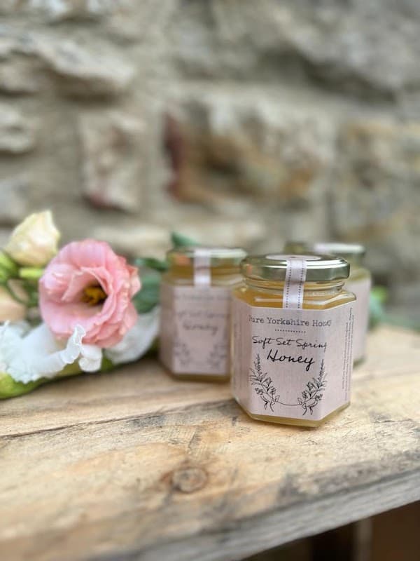 Three jars of Pure Yorkshire Honey on a wooden surface, surrounded by soft pink flowers.