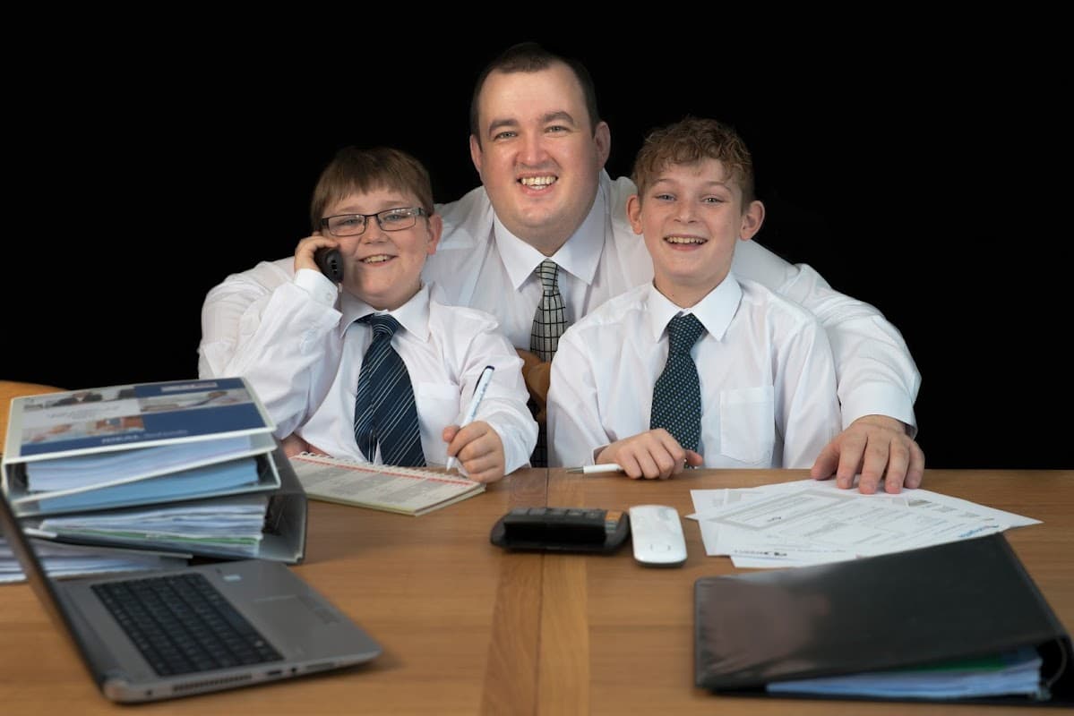 A man and two boys in white shirts and ties sit at a desk with paperwork and a laptop, smiling at the camera.