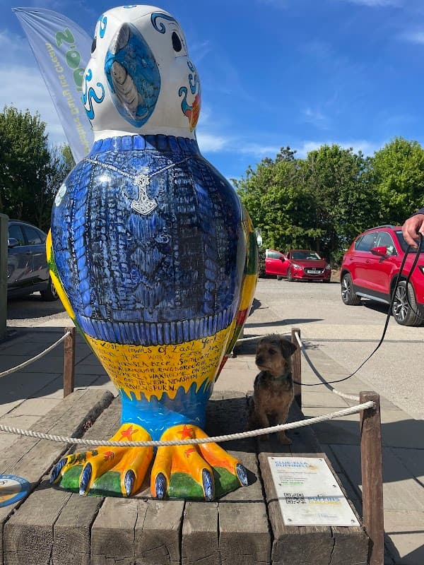 Colorful bird sculpture with intricate designs, a small dog sitting nearby, and parked cars in the background.