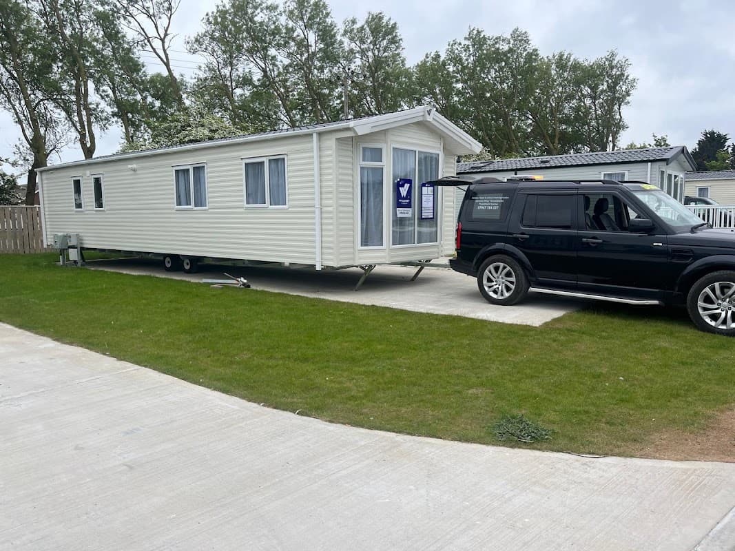 A static caravan parked on a concrete pad with a black SUV nearby, surrounded by green grass and trees.