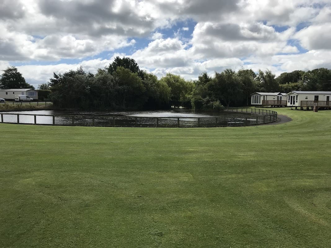 A serene caravan park with a pond, surrounded by greenery and holiday homes under a cloudy sky.