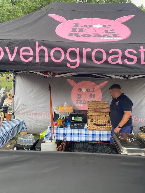 Market stall with a black canopy, blue checkered tablecloth, and a vendor preparing food. Sign reads "Love Hog Roast."
