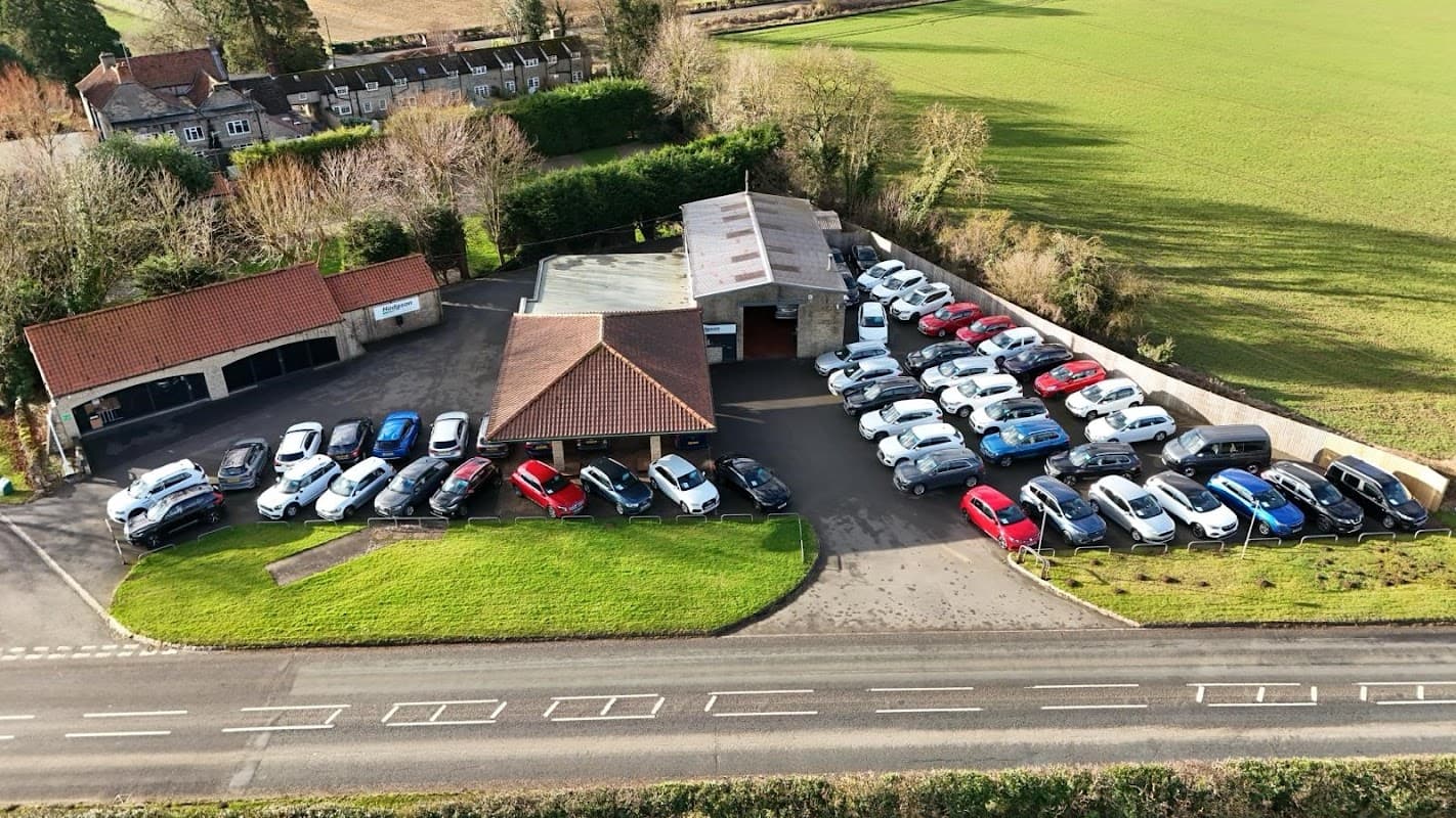 Hodgson Car Sales with a variety of parked cars, surrounded by green fields and a residential area in Snainton, Yorkshire.