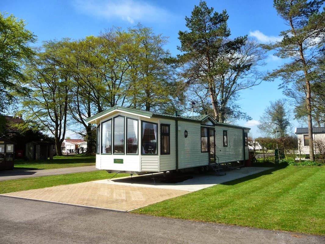 Caravan parked on a paved area, surrounded by green grass and trees under a clear blue sky.