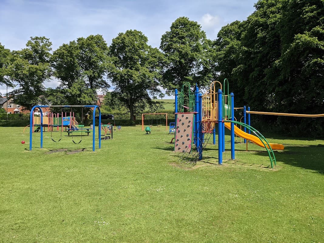 Colorful playground equipment with slides, climbing walls, and swings on a grassy area surrounded by trees.