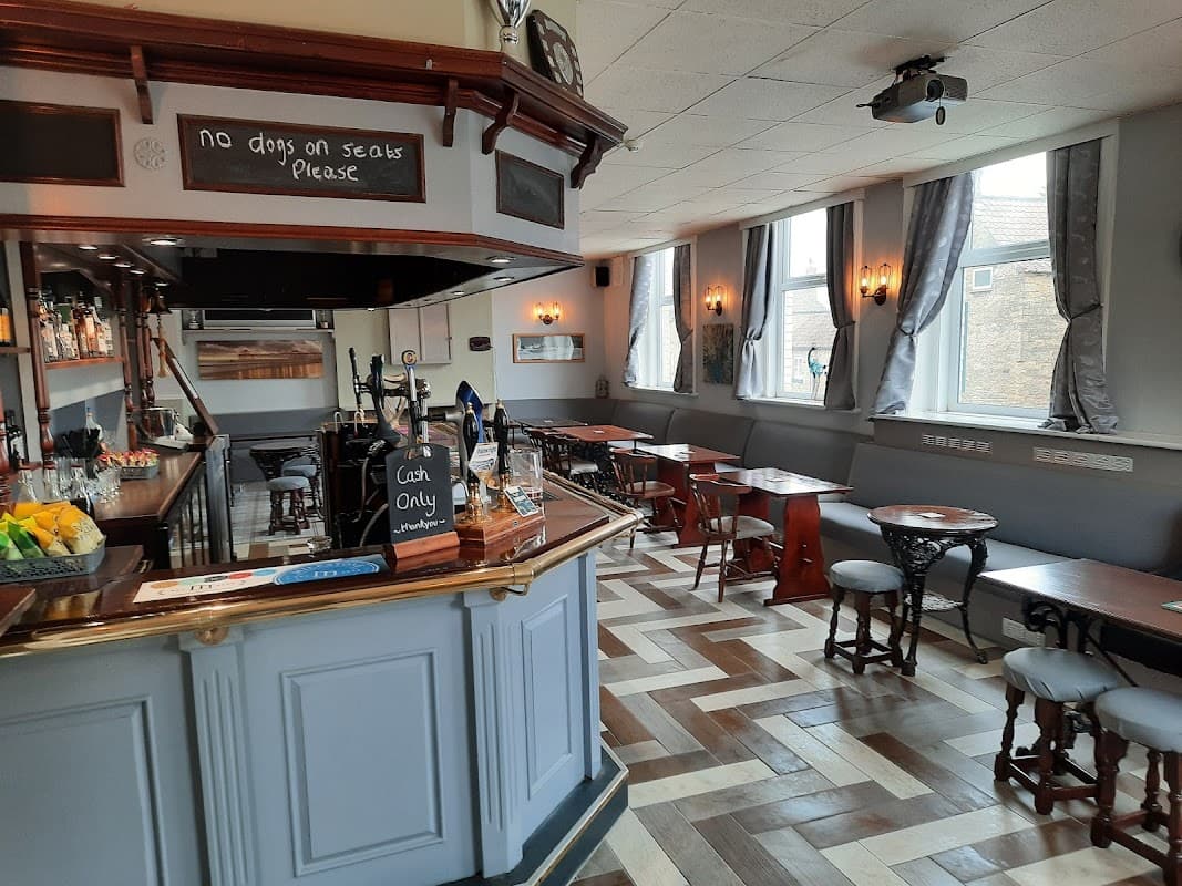 Cozy bar interior with wooden furniture, a polished counter, and large windows, featuring a no dogs sign.