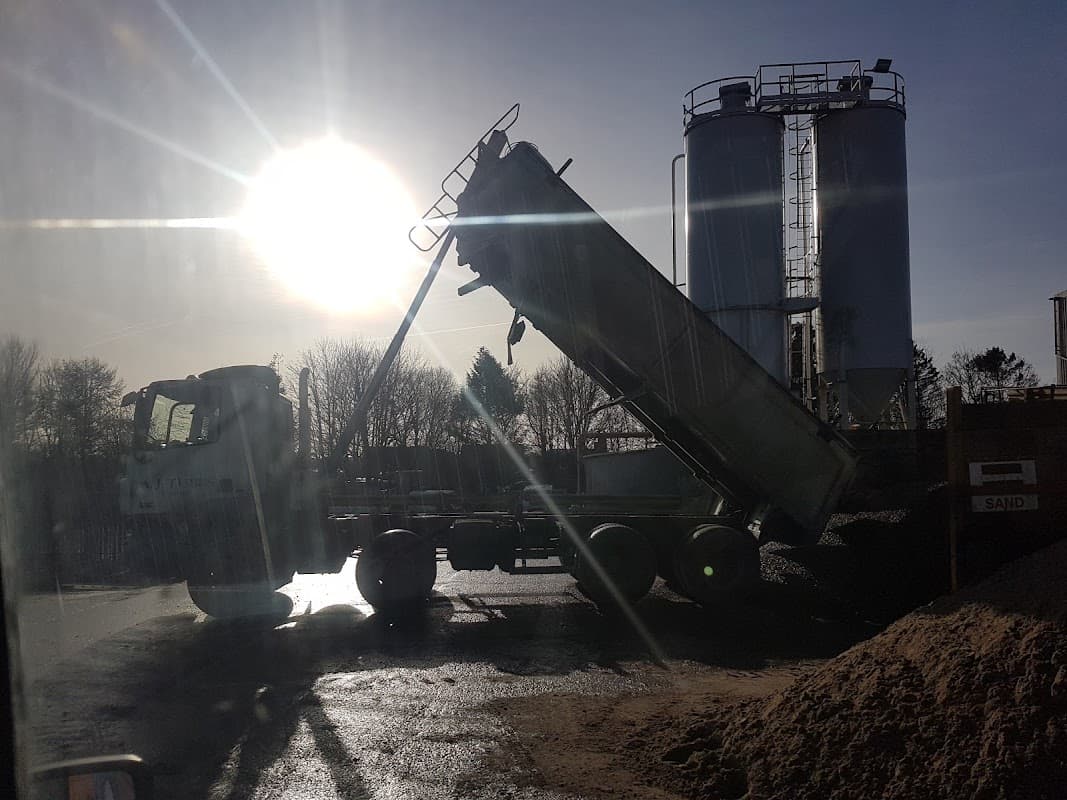 A truck unloading material beside large silos, with sunlight shining in the background and a sandy pile nearby.
