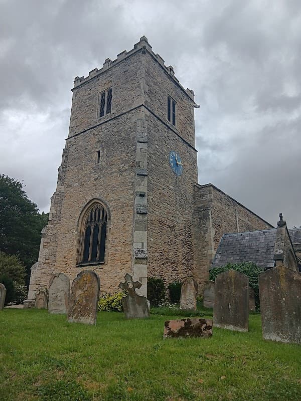 Historic stone church tower with a blue clock, surrounded by gravestones and greenery under a cloudy sky.