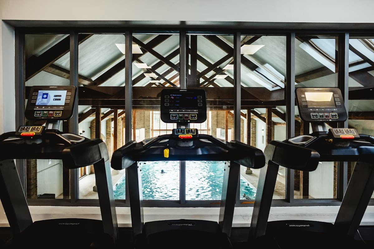Treadmills with screens facing a swimming pool, framed by large windows and a wooden-beamed ceiling.