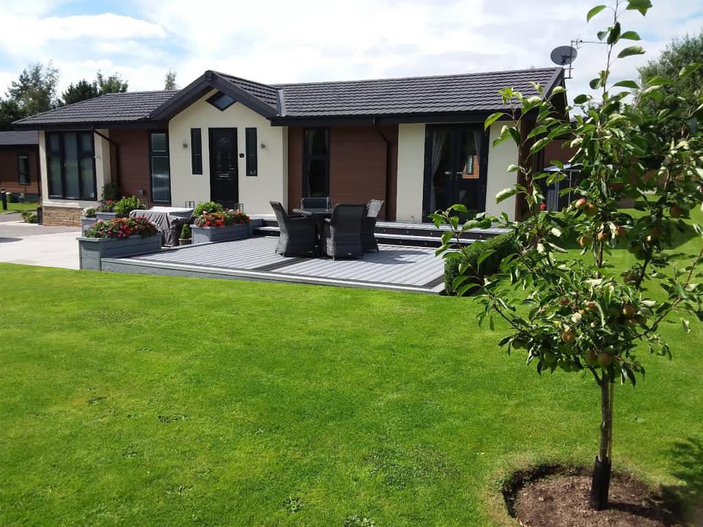 Modern bungalow with a wooden deck, outdoor seating, and landscaped lawn, surrounded by greenery at Thornborough Grange Park.