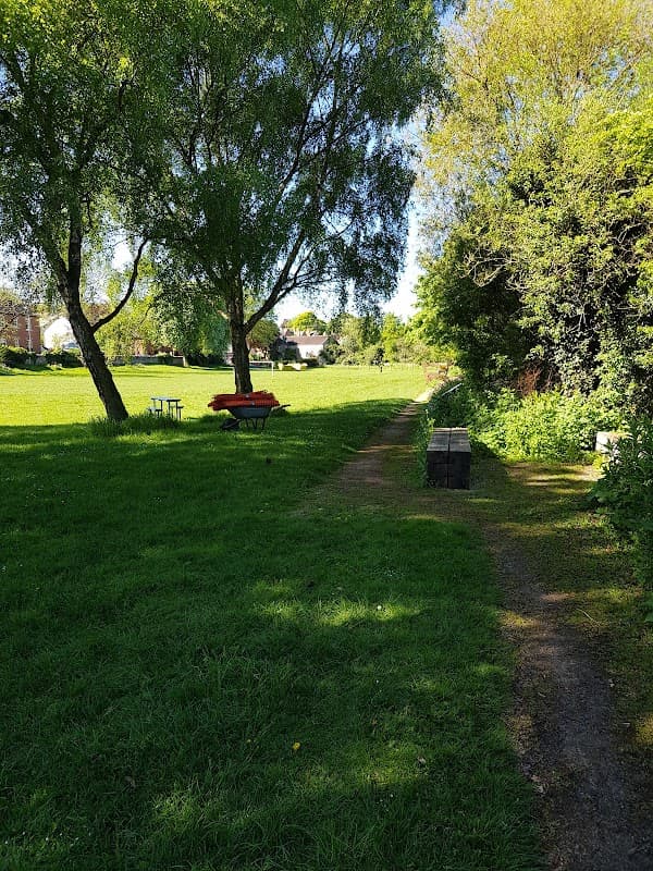 Lush green park with trees, a pathway, benches, and a picnic table under clear blue skies in South Milford.