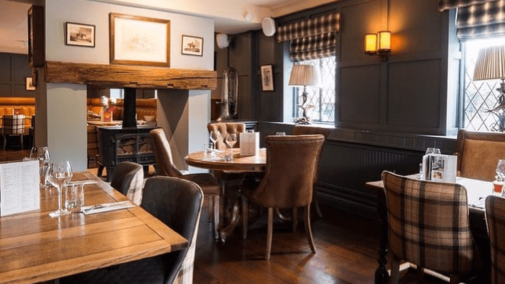 Cozy restaurant interior with wooden tables, plush chairs, and warm lighting, featuring a fireplace and framed artwork.
