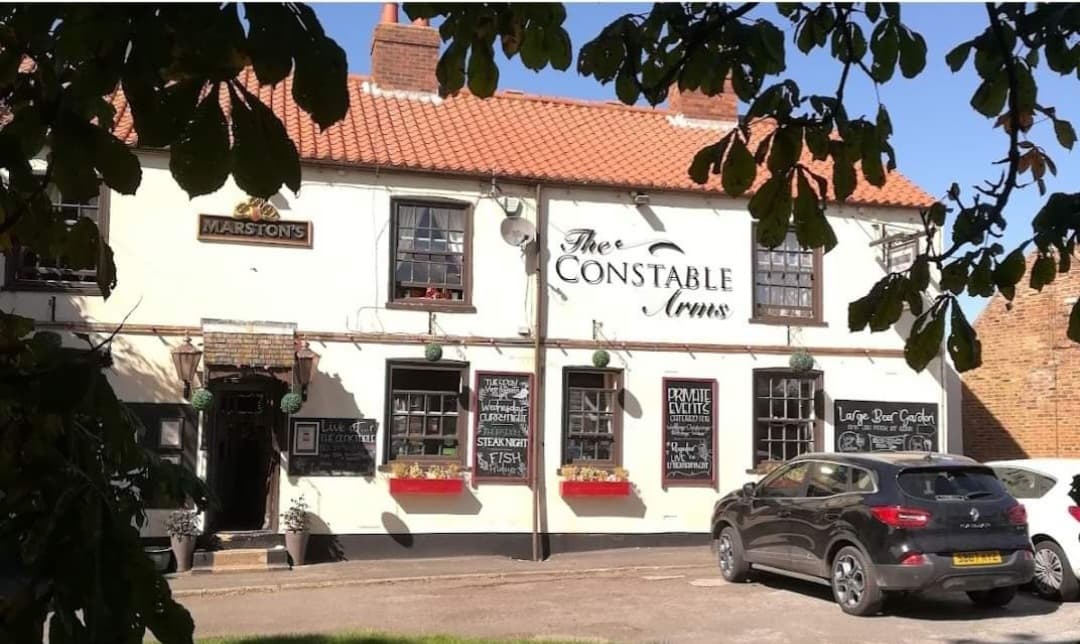 Traditional pub with a white exterior, red roof, and black signage, surrounded by greenery and parked cars.