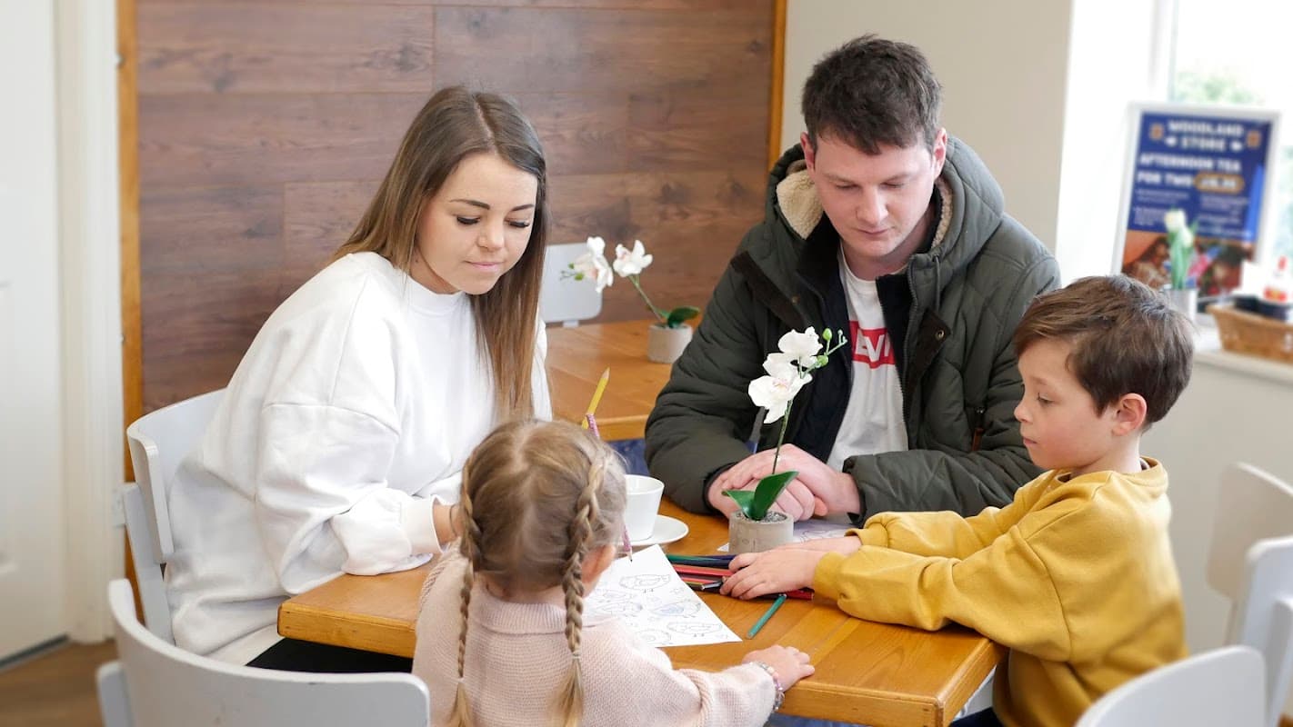 A family at a wooden table, coloring with crayons, surrounded by a cozy interior and a small potted plant.
