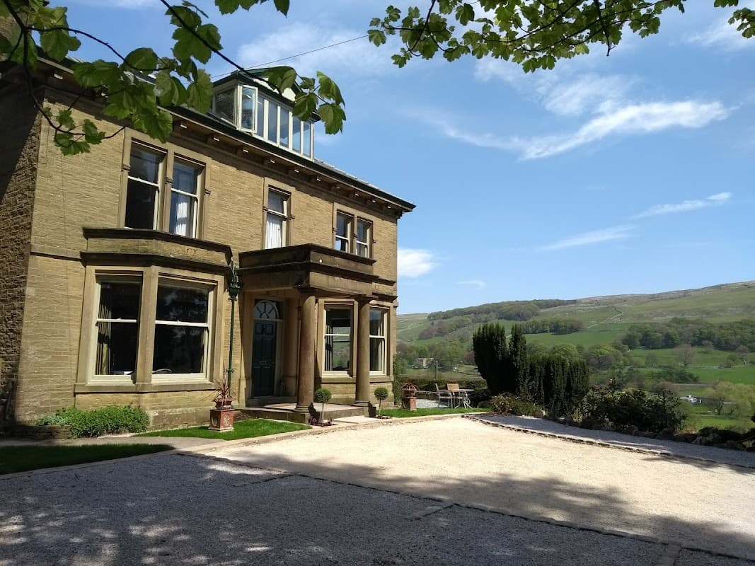 Victorian-style house with large windows, surrounded by greenery and hills under a clear blue sky.