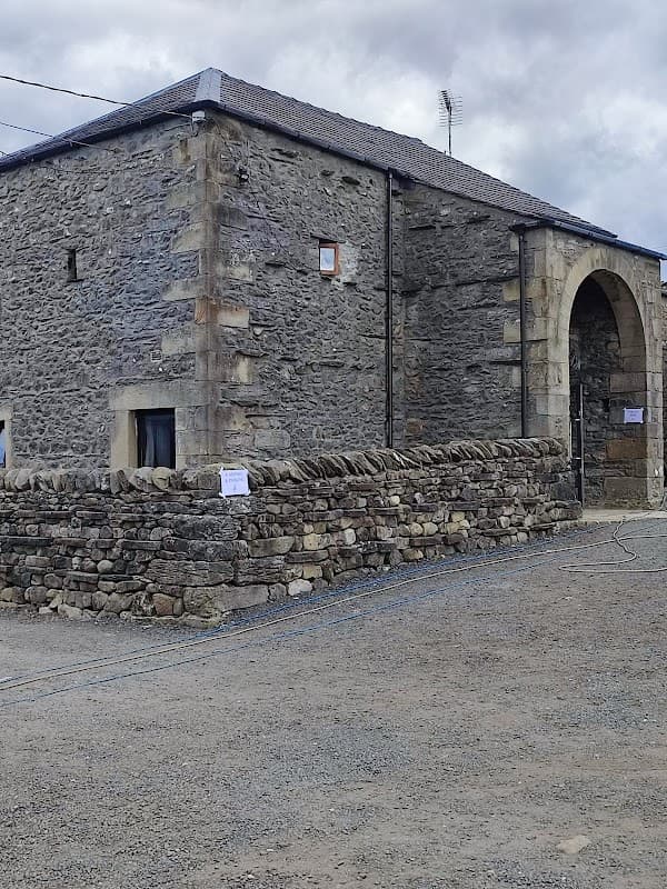 Stone barn with a large archway entrance, surrounded by a low stone wall and gravel path, set against a cloudy sky.