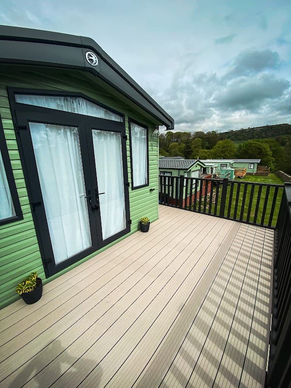 Green cabin with large glass doors on a deck, overlooking grassy areas and other cabins under a cloudy sky.